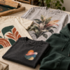 Printed T-shirts and an embroidered green hoodie beside a screen-printing frame, thread spools and embroidery hoop.